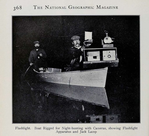 Photo of George Shiras III in a boat rigged for night-hunting with cameras, showing flashlight apparatus and jack lamp.